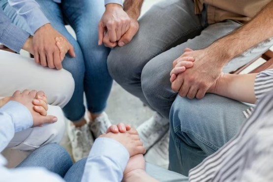 A imagem mostra um grupo de pessoas sentadas em círculo, segurando as mãos umas das outras. As mãos conectadas transmitem uma sensação de união e apoio mútuo, sugerindo que o grupo está em um momento de solidariedade, talvez em uma reunião de suporte emocional ou espiritual. A foto foca nas mãos e pernas dos participantes, com roupas casuais, dando uma impressão de intimidade e proximidade entre eles.