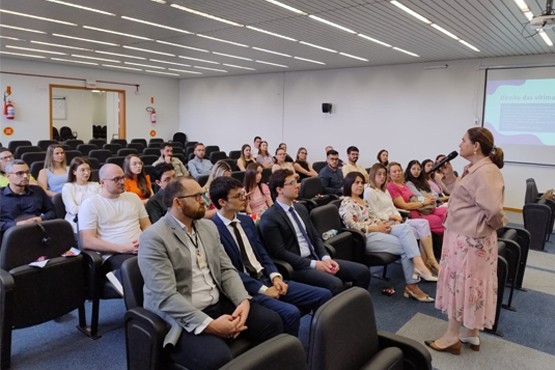 A imagem mostra uma sala de conferências com várias pessoas sentadas em cadeiras, prestando atenção a uma apresentação. À direita, uma mulher em pé, segurando um microfone, está falando para o público. A apresentação parece ser formal, pois muitos dos participantes estão vestidos de forma profissional, com trajes de negócios, especialmente aqueles sentados nas primeiras fileiras. No fundo, há uma tela de projeção onde uma apresentação está sendo exibida, e parte de um slide pode ser visto com o título "Direito das vítimas", sugerindo que o evento ou palestra é relacionado a temas jurídicos ou de direitos humanos, com foco na proteção das vítimas. O público está atento, composto por homens e mulheres de diferentes idades, em um ambiente que parece ser acadêmico ou institucional. A atmosfera da sala é de concentração e engajamento, e o evento parece ser de natureza educacional ou de conscientização.