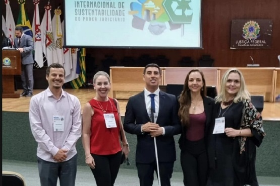 A imagem mostra um grupo de cinco pessoas posando para uma foto em um ambiente formal, possivelmente um auditório ou sala de conferências. Ao fundo, há um púlpito e várias bandeiras. As pessoas estão vestidas de maneira formal. No canto direito da imagem, há uma tela de projeção com um logotipo e texto que diz "INTERNACIONAL DE SUSTENTABILIDADE DO PODER JUDICIÁRIO". A imagem está ligeiramente inclinada para a esquerda.