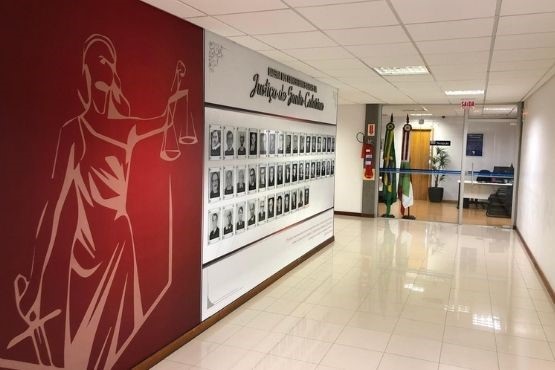 A imagem mostra um corredor com paredes revestidas de azulejos claros. No teto, há uma grande ilustração em vermelho e branco de uma figura estilizada segurando uma balança, que é um símbolo comum da justiça. Na parede à direita, há uma fileira de retratos em preto e branco, provavelmente de pessoas importantes ou funcionários, com o título "Juízes" ao lado. Ao fundo do corredor, há uma porta de vidro com algumas plantas visíveis atrás dela.