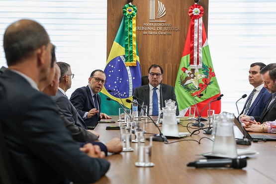 A imagem mostra uma reunião em uma sala de conferências. Há várias pessoas sentadas ao redor de uma mesa de madeira. Na mesa, há garrafas de água, copos e papéis. As pessoas estão vestidas formalmente. Na parede ao fundo, há duas bandeiras. A sala parece bem iluminada e as pessoas estão atentas à reunião