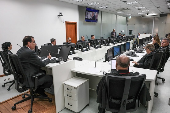 Foto horizontal: Francisco Oliveira Neto participa de sessão colegiada, magistrados em bancada com computadores, ambiente do tribunal.