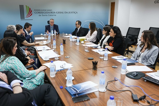 A imagem mostra uma reunião formal em uma sala de conferências, com várias pessoas sentadas ao redor de uma mesa de madeira grande. A mesa está coberta com garrafas de água, papéis, cadernos e alguns dispositivos eletrônicos, como um microfone central. No fundo, há um painel azul com o texto "Poder Judiciário Corregedoria-Geral da Justiça de Santa Catarina" e o logotipo correspondente. Uma pessoa, que parece ser a figura principal ou o presidente da reunião, está falando enquanto os demais participantes, vestidos de forma profissional, prestam atenção. A cena transmite um ambiente institucional de discussão ou planejamento dentro do Judiciário.