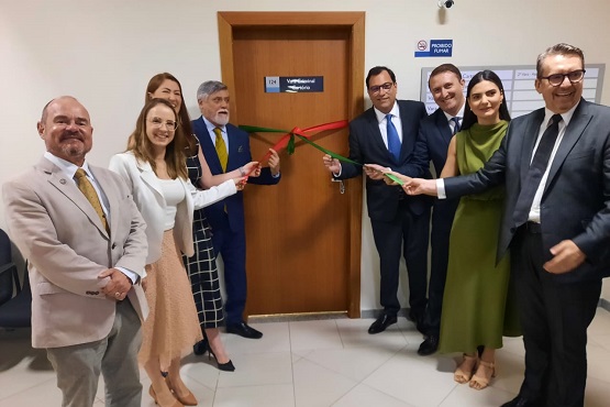 A imagem mostra uma cerimônia de inauguração, onde um grupo de pessoas está reunido em frente a uma porta, segurando uma fita nas cores verde e vermelha prestes a ser cortada. Todos estão vestidos formalmente, e alguns sorriem para a câmera. A placa na porta indica que se trata de um ambiente oficial ou institucional, e o evento parece ser uma inauguração ou abertura de um novo espaço ou serviço. A atmosfera é de celebração e solenidade, com a presença de autoridades ou representantes envolvidos na ocasião.