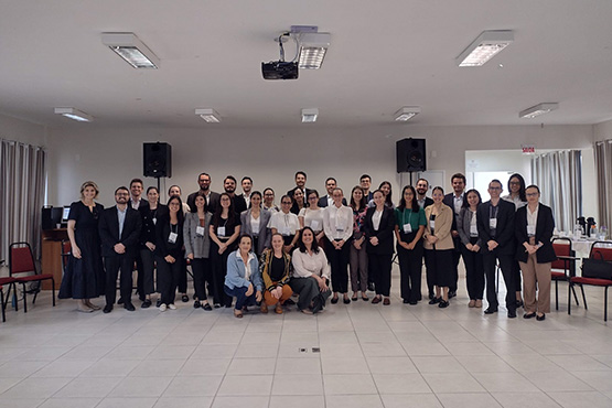 A foto mostra um grupo grande de pessoas posando juntas em um ambiente fechado. A maioria está vestida formalmente, sugerindo que pode se tratar de um evento acadêmico, uma conferência, ou uma reunião profissional. O ambiente é uma sala grande, com paredes claras, um teto com luminárias, e alguns equipamentos de áudio ao fundo. Há cadeiras vermelhas ao redor da sala, e todos os presentes na foto parecem estar em clima amigável, com sorrisos.