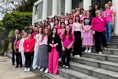 A foto mostra um grupo grande de pessoas posando em uma escadaria, em frente a um prédio com colunas e detalhes arquitetônicos clássicos. Muitas pessoas na imagem estão vestindo roupas em diferentes tons de rosa, sugerindo que estão participando de uma campanha de conscientização, como o Outubro Rosa, que promove a prevenção ao câncer de mama. O grupo é composto por uma mistura de pessoas de diferentes idades, e o clima é alegre e acolhedor, com muitos sorrisos. A vegetação ao fundo contribui para um cenário harmonioso.