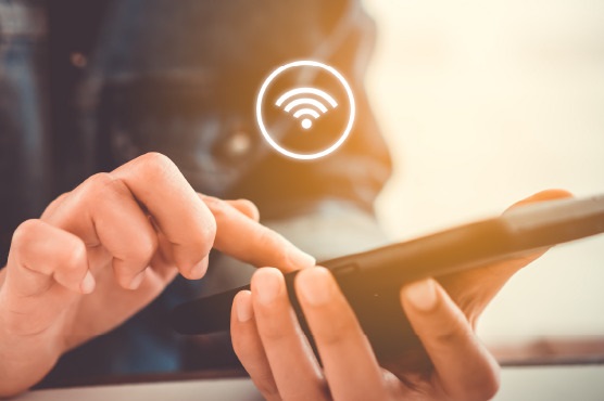 A imagem mostra uma pessoa usando um dispositivo móvel, provavelmente um smartphone, com as mãos focadas na tela. Há um ícone flutuante de sinal de Wi-Fi desenhado acima, indicando a conexão ou o uso de uma rede sem fio. O fundo da imagem é suavemente desfocado, com uma iluminação quente e acolhedora.