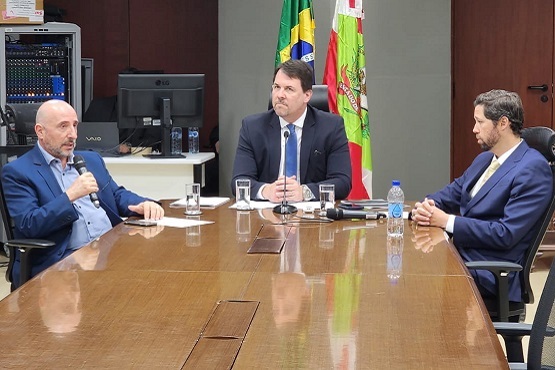 A imagem mostra três homens sentados ao redor de uma grande mesa de madeira em uma sala formal. Dois deles estão vestidos com ternos escuros, e o terceiro, à esquerda, também está vestido de forma elegante, segurando um microfone enquanto fala. No fundo, há bandeiras do Brasil e de Santa Catarina, sugerindo um ambiente institucional ou governamental. A mesa tem microfones e copos d'água, o que indica que pode estar ocorrendo uma reunião oficial ou coletiva de imprensa.