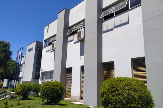 A imagem mostra a fachada de um prédio de dois andares com um design moderno e linhas retas. O prédio é pintado de branco e cinza, e possui várias janelas com toldos abertos. Há aparelhos de ar-condicionado instalados em algumas janelas. Na frente do prédio, há arbustos bem cuidados e uma área gramada. À esquerda da imagem, há três mastros com bandeiras hasteadas. O céu está azul, indicando um dia ensolarado