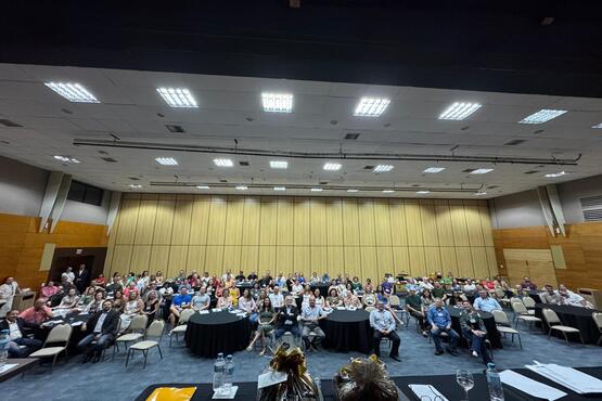 A imagem mostra uma sala de conferências grande, com muitas pessoas sentadas em cadeiras organizadas em fileiras. A sala tem um teto alto com várias luzes embutidas, e as paredes são revestidas com painéis de madeira clara. Na frente, há algumas mesas redondas cobertas com toalhas pretas, e parece haver alguns objetos decorativos ou presentes sobre elas. As pessoas na audiência estão olhando para a câmera, e a sala está bem iluminada. Parece ser um evento ou uma apresentação, dado o número de pessoas presentes e a disposição do espaço.