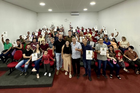 A imagem mostra um grupo grande de pessoas em um auditório. Elas estão sentadas em cadeiras vermelhas e muitas delas estão segurando certificados ou diplomas. Na frente do grupo, há algumas pessoas em pé, sorrindo para a câmera. O ambiente é iluminado e as paredes são brancas. Parece ser uma cerimônia de entrega de certificados ou diplomas. Há uma mistura de homens, mulheres e crianças na foto, todos parecendo felizes e orgulhosos.