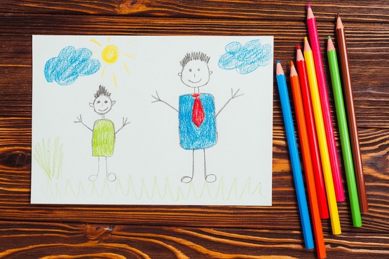 A imagem mostra um desenho infantil feito com lápis de cor. O desenho está em uma folha de papel sobre uma superfície de madeira. Nele, há duas figuras humanas desenhadas de forma simples. A figura à esquerda é menor, com cabelo curto e preto, vestindo uma roupa verde. A figura à direita é maior, também com cabelo curto e preto, vestindo uma roupa azul com uma gravata vermelha. Acima das figuras, há duas nuvens azuis e um sol amarelo. No canto inferior esquerdo, há algumas linhas verdes que parecem representar grama. Ao lado direito do desenho, há um conjunto de lápis de cor em várias cores: vermelho, rosa, laranja, verde, azul, amarelo e marrom.