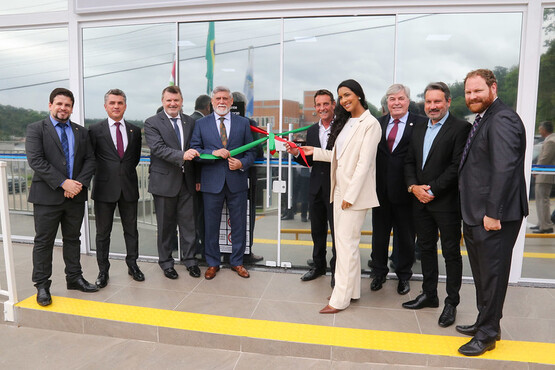 A imagem mostra um grupo de nove pessoas em frente a um prédio com fachada de vidro. No centro, duas pessoas estão segurando uma fita verde e vermelha, prestes a cortá-la, sugerindo uma cerimônia de inauguração. A pessoa à esquerda que segura a fita é um homem de terno azul escuro e barba grisalha. A pessoa à direita é uma mulher vestida com um terno branco e cabelo escuro preso. As outras pessoas ao redor estão vestidas formalmente, com ternos e gravatas. Ao fundo, é possível ver bandeiras hasteadas e um céu nublado.