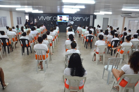 A imagem mostra uma sala ampla com várias pessoas sentadas em cadeiras de plástico, organizadas em fileiras. As pessoas estão vestindo uniformes brancos com detalhes laranja, que parecem ser roupas de detentos. Na frente da sala, há um palco com uma tela grande exibindo uma imagem. Acima da tela, há uma frase escrita na parede: "viver, amar e servir". A sala é bem iluminada, com luzes fluorescentes no teto. As cadeiras são de cores variadas, principalmente brancas e pretas.