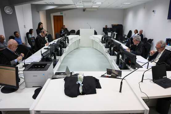 A imagem mostra uma sala de tribunal moderna, com uma mesa em formato de "U" onde várias pessoas estão sentadas, possivelmente advogados e juízes. Há computadores e microfones sobre a mesa. No centro da mesa, há um manto preto dobrado com um martelo de juiz em cima. Ao fundo, há um púlpito com um microfone. As paredes são brancas e o ambiente é bem iluminado, com luzes no teto. Algumas pessoas estão sentadas ao longo das paredes, observando.