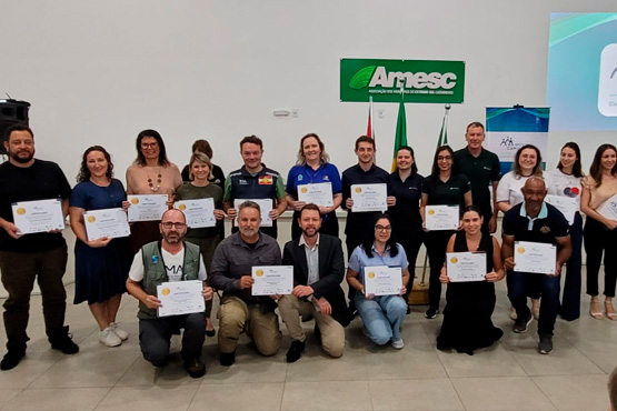 A imagem mostra um grupo de pessoas posando para uma foto em um ambiente interno. Ao todo, há 20 pessoas na imagem, distribuídas em duas fileiras. A maioria delas está segurando certificados, sugerindo que participaram de algum tipo de cerimônia de premiação ou conclusão de curso. No fundo, há um banner verde com o logotipo da "Amesc" e algumas bandeiras. As pessoas estão sorrindo e parecem felizes. O ambiente é bem iluminado e tem um piso claro.