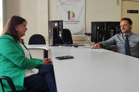 A imagem mostra duas pessoas sentadas em lados opostos de uma mesa em um escritório. À esquerda, uma mulher de cabelos castanhos veste um blazer verde e está sentada de perfil. À direita, um homem de cabelos curtos e escuros veste uma camisa cinza com gravata e está olhando para a mulher. Na mesa, há um celular próximo à mulher e alguns papéis próximos ao homem. Ao fundo, há dois monitores de computador e um tripé pequeno. Na parede, há um banner com o logotipo e o texto "Poder Judiciário de Santa Catarina".