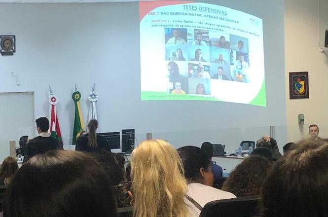 A imagem mostra uma sala de aula ou auditório com várias pessoas sentadas, olhando para uma apresentação projetada em uma tela na frente da sala. Na tela, há uma apresentação com o título "TESES DEFENSIVAS" e o subtítulo "MENTIRA 3: NÃO QUERIAM MATAR, APENAS MACHUCAR". Abaixo do subtítulo, há uma lista de nomes e fotos de pessoas. A sala tem bandeiras do Brasil e de outro estado ou município ao lado da tela. As pessoas na sala estão prestando atenção na apresentação.