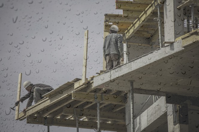 A imagem mostra dois trabalhadores da construção civil em um prédio em construção. Eles estão usando capacetes de segurança e roupas de trabalho. Um dos trabalhadores está agachado, aparentemente ajustando ou fixando uma estrutura de madeira, enquanto o outro está em pé, olhando para a frente. A estrutura em que eles estão trabalhando é feita de concreto e madeira, com andaimes de metal visíveis. A imagem parece ter sido tirada através de uma janela com gotas de chuva, pois há marcas de gotas de água espalhadas por toda a imagem. O céu está nublado, sugerindo um dia chuvoso.