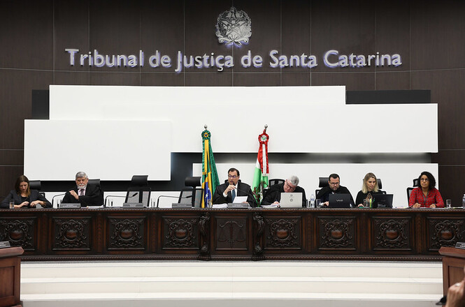 A imagem mostra uma sala de tribunal com uma mesa comprida onde estão sentadas sete pessoas. Acima delas, está escrito "Tribunal de Justiça de Santa Catarina" em letras grandes. Na parede, há um brasão acima do texto. Na mesa, há dois mastros com bandeiras: uma do Brasil e outra do estado de Santa Catarina. As pessoas na mesa parecem estar participando de uma sessão ou reunião oficial, com papéis, laptops e microfones à sua frente. O ambiente é formal e bem iluminado.