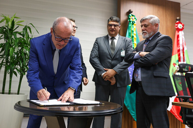 A imagem mostra quatro homens em um ambiente formal, provavelmente um escritório ou sala de reuniões. O homem à esquerda está assinando um documento sobre uma mesa redonda. Ele veste um terno azul e gravata cinza. Atrás dele, há três homens de pé, observando. Todos estão vestidos com ternos escuros. No fundo, há duas bandeiras, uma do Brasil e outra que parece ser de um estado brasileiro, possivelmente Santa Catarina, devido às cores e ao brasão visíveis. À esquerda, há uma planta em um vaso. O ambiente parece ser formal e oficial, sugerindo que a assinatura pode ser de um documento importante.