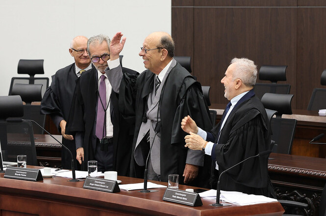 A imagem mostra quatro homens vestidos com togas, aparentemente em uma cerimônia ou sessão formal em um tribunal ou órgão judicial. Eles estão de pé em volta de uma mesa com microfones, copos de água e placas de identificação. Eles parecem estar conversando e rindo de maneira descontraída, o que cria um clima informal e amistoso entre eles. O ambiente é formal, com móveis de madeira escura e cadeiras de design sóbrio ao fundo, sugerindo um cenário oficial, possivelmente de uma sessão de julgamento ou reunião de magistrados.