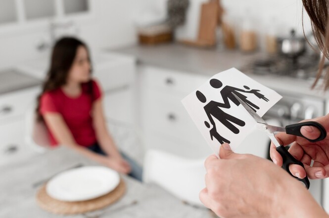 A imagem mostra uma cena em uma cozinha. Em primeiro plano, há uma pessoa segurando uma tesoura e cortando uma imagem de papel que representa uma família estilizada, composta por figuras de um adulto e duas crianças. Ao fundo, desfocada, há uma menina sentada à mesa, vestindo uma camiseta vermelha. A mesa tem um prato branco sobre um jogo americano. A cozinha é moderna, com armários brancos e uma bancada de mármore. A cena sugere um momento de tensão ou simbolismo, possivelmente relacionado a questões familiares.