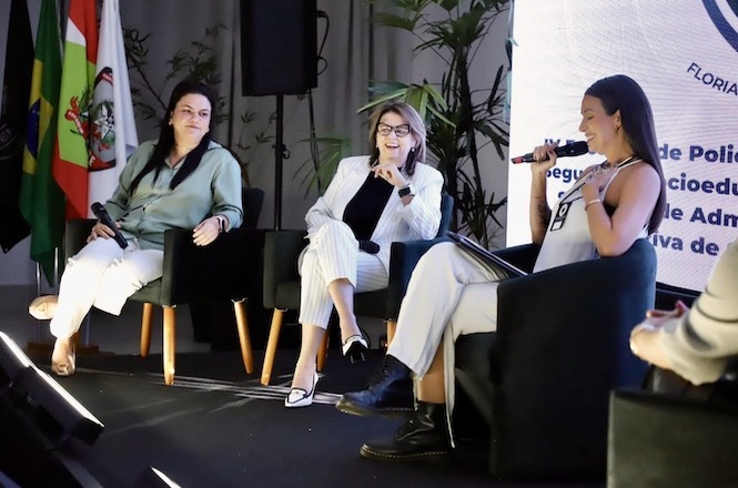 A imagem mostra três mulheres sentadas em cadeiras em um palco, participando de um painel ou discussão em um ambiente formal, como uma conferência ou seminário. Todas estão segurando microfones, sugerindo que estão envolvidas em uma conversa ou debate. A mulher à direita está falando, enquanto as outras duas escutam com atenção. No fundo, há bandeiras e plantas decorativas, e o logotipo do evento pode ser visto parcialmente. As roupas das participantes são formais e elegantes, e a atmosfera geral é de profissionalismo e seriedade, com uma iluminação focada no palco.