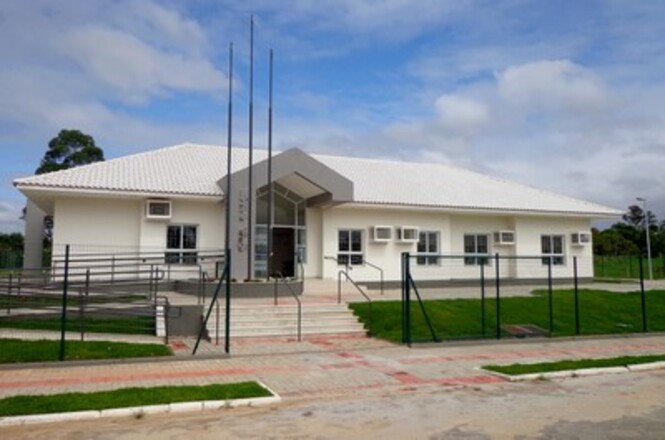 A imagem mostra um edifício de um andar com um design simples e moderno, com paredes claras e telhado inclinado. A entrada principal tem uma estrutura triangular em destaque, e há uma rampa de acessibilidade ao lado da escada. Na frente, há três mastros para bandeiras, indicando um possível prédio público ou institucional. O edifício está cercado por uma grade baixa e há uma área gramada ao redor, além de uma calçada pavimentada na frente. O céu está parcialmente nublado, proporcionando uma iluminação suave ao local.