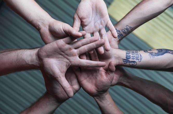 A imagem mostra várias mãos unidas em um gesto de união e colaboração, formando um círculo com as palmas voltadas para baixo. Algumas das mãos possuem tatuagens visíveis, indicando diversidade entre os participantes. A disposição das mãos sugere um simbolismo de solidariedade, trabalho em equipe e cooperação. O fundo é desfocado, com tons neutros, de forma que o foco principal está nas mãos e na mensagem de unidade que elas transmitem.