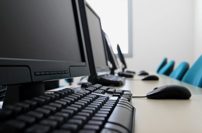 A imagem mostra uma fileira de computadores em uma sala de informática ou laboratório de tecnologia. Cada estação de trabalho possui um monitor, teclado e mouse alinhados sobre uma mesa, indicando um ambiente organizado e preparado para atividades de ensino, treinamento ou trabalho colaborativo. As cadeiras azuis ao fundo sugerem um ambiente institucional, possivelmente uma escola, universidade ou escritório. A iluminação é suave e o ambiente parece limpo e bem conservado, adequado para um espaço de aprendizado ou trabalho.