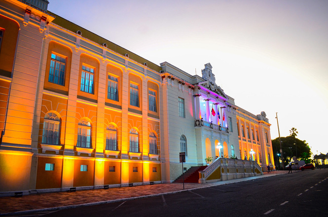 A imagem mostra um edifício histórico e imponente, iluminado por luzes coloridas que destacam sua arquitetura clássica. A fachada é detalhada, com janelas em arco e ornamentos decorativos, e a iluminação em tons de laranja e roxo cria um efeito visual impressionante contra o céu ao entardecer. Uma escadaria leva à entrada principal, onde há bandeiras hasteadas, sugerindo que o prédio possui importância cívica ou institucional, possivelmente uma prefeitura, tribunal ou museu. O cenário transmite uma atmosfera de respeito e tradição, destacando o valor cultural e arquitetônico do edifício.