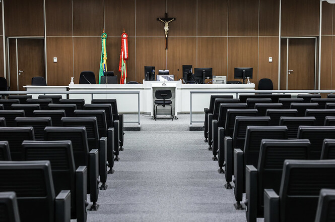 A imagem mostra o interior de uma sala de audiências ou tribunal. No centro, há uma bancada elevada onde ficam o juiz, advogados e outros oficiais de justiça, com cadeiras voltadas para a frente. Acima da bancada, há um crucifixo fixado na parede, e ao lado direito do crucifixo, bandeiras do Brasil e de outro estado ou instituição. No fundo da sala, há três monitores sobre a bancada, indicando que a tecnologia pode ser utilizada nas sessões. A sala é composta por várias fileiras de cadeiras em preto para o público, dispostas em linhas simétricas, com corredores nas laterais e no centro. A decoração é sóbria, com tons neutros e materiais que dão um ar formal ao ambiente.