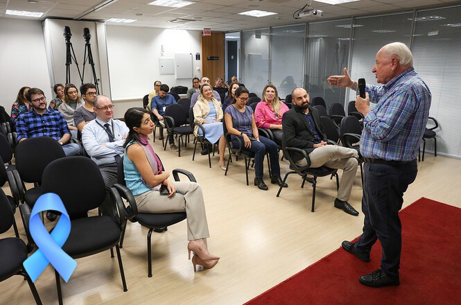 A imagem mostra uma palestra ou reunião em uma sala de conferências. Um homem mais velho, segurando um microfone, está de pé na frente da sala, falando para o público. Ele gesticula com a mão, indicando que está envolvido em sua fala. Os participantes estão sentados em cadeiras pretas, prestando atenção ao palestrante, com alguns sorrindo ou observando atentamente. No canto da imagem, há um laço azul, possivelmente indicando apoio a uma campanha, como a de conscientização sobre o câncer de próstata (Novembro Azul). A sala tem um ambiente profissional, com piso de madeira clara e uma parede de vidro ao fundo.
