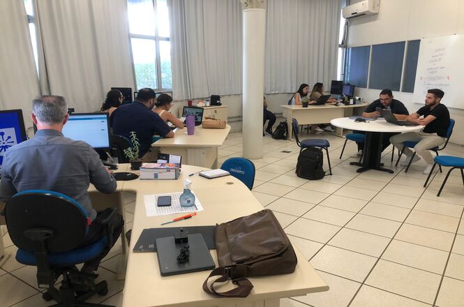 A imagem mostra uma sala de estudos ou um espaço de trabalho colaborativo. Há várias pessoas sentadas em mesas brancas, utilizando laptops e computadores. Algumas pessoas trabalham individualmente, enquanto outras parecem estar em grupo, interagindo entre si. A sala é iluminada, com janelas grandes ao fundo, cobertas por cortinas. As cadeiras são azuis, e há alguns objetos pessoais, como garrafas de água, mochilas e cadernos sobre as mesas. No canto direito da imagem, há um quadro branco com anotações e, acima dele, um aparelho de ar-condicionado. O ambiente transmite uma atmosfera de foco e colaboração, sugerindo um espaço de ensino ou de trabalho acadêmico.