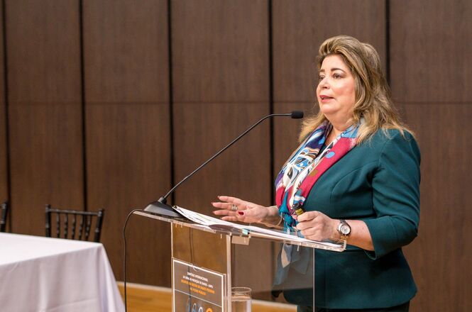 A imagem mostra uma mulher falando em um púlpito, aparentemente em um evento formal ou cerimônia. Ela usa um blazer verde e um lenço colorido em volta do pescoço. A expressão dela é séria e focada, indicando que está envolvida em seu discurso. Sobre o púlpito de acrílico transparente, há um microfone e alguns papéis, sugerindo que ela pode estar lendo ou consultando notas. O fundo é composto por uma parede de madeira escura, e ao lado do púlpito há uma mesa coberta com uma toalha branca. A imagem transmite uma sensação de profissionalismo e solenidade, indicando que a mulher tem uma posição de destaque no evento.