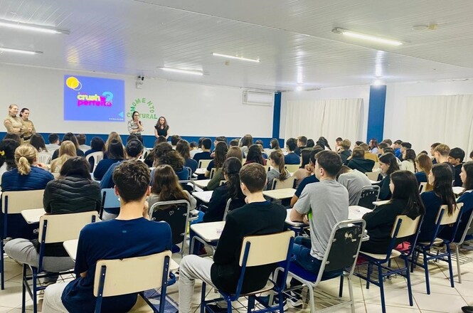 A imagem mostra uma sala de aula ou auditório com várias fileiras de cadeiras ocupadas por estudantes, todos voltados para a frente, onde três mulheres estão em pé, provavelmente apresentando algo para o grupo. Na parede frontal, há uma projeção que exibe o título "Crush Perfeito" e uma imagem relacionada, sugerindo que é o tema da apresentação. A sala é espaçosa, com paredes brancas e janelas cobertas por cortinas. O ambiente é iluminado por luzes fluorescentes no teto. A presença de estudantes atentos indica que o evento é voltado para aprendizado ou orientação, possivelmente um workshop, palestra ou aula interativa.