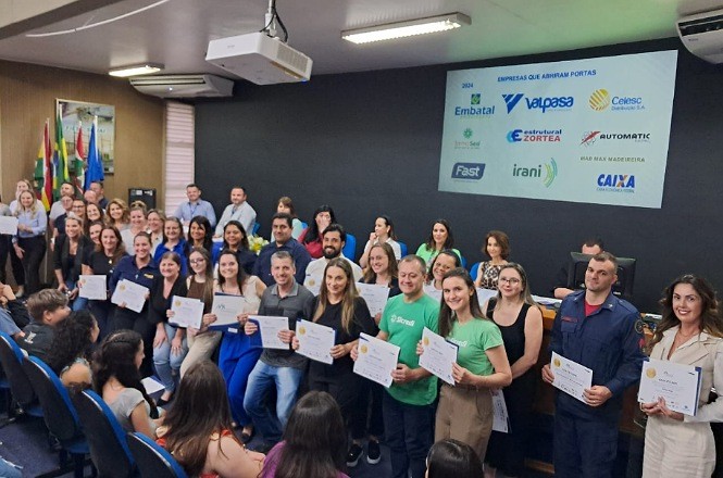 A imagem mostra um grupo de pessoas em um auditório, posando para uma foto coletiva enquanto seguram certificados. Esse momento parece ser uma cerimônia de formatura, entrega de certificados ou reconhecimento, com todos sorrindo para a câmera. Ao fundo, há um painel com logotipos de diversas empresas, indicando que são "Empresas que abrem portas," o que sugere uma parceria ou apoio dessas empresas ao evento ou programa. Bandeiras estão posicionadas no lado esquerdo da imagem, contribuindo para a formalidade do ambiente. A cena reflete um clima de celebração e conquista, com os participantes comemorando suas realizações.