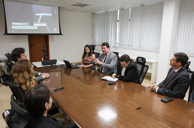 A imagem mostra uma reunião formal em uma sala de conferências. Há sete pessoas sentadas ao redor de uma mesa retangular de madeira. No centro, uma pessoa gesticula enquanto fala, sugerindo que está liderando a discussão. Todos estão vestidos formalmente, a maioria em trajes de trabalho, incluindo ternos e camisas sociais. Na parede ao fundo, há uma tela de projeção que exibe uma apresentação com o título "FORMAÇÃO INICIAL PARA SERVIDORES E GESTORES", indicando que o tema da reunião pode estar relacionado ao treinamento ou capacitação. A sala possui persianas cobrindo as janelas, e é bem iluminada, com luzes de teto brancas. Os participantes parecem atentos, alguns tomando notas, enquanto outros observam o orador. A atmosfera parece séria e profissional, sugerindo uma reunião administrativa ou de planejamento.