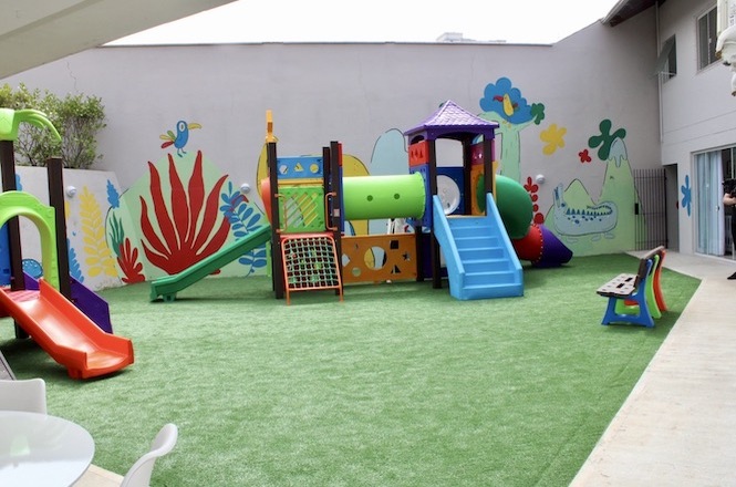 A imagem mostra um playground infantil ao ar livre, equipado com brinquedos coloridos sobre um piso de grama sintética verde. Entre os brinquedos, há escorregadores, túneis, uma pequena parede de escalada e um conjunto de estruturas para escalar e brincar. As paredes ao redor do espaço são decoradas com desenhos vibrantes de plantas, flores e animais, criando um ambiente alegre e estimulante para as crianças. Há também alguns bancos e cadeiras, indicando áreas para descanso ou supervisão. O espaço parece seguro e bem cuidado, ideal para atividades recreativas infantis.