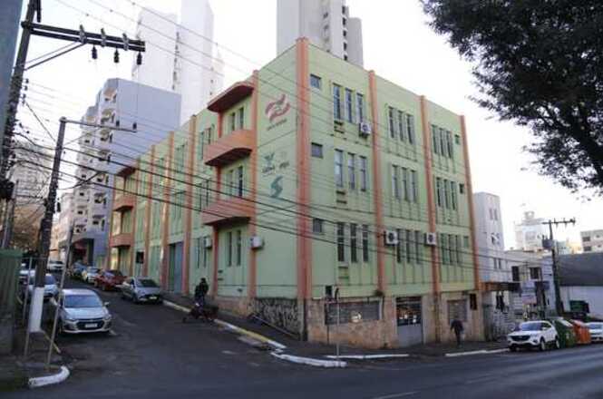 A imagem mostra um prédio de três andares em uma esquina de rua urbana. O edifício possui fachada em tom verde claro com detalhes em cor laranja nas bordas e divisões das janelas. Algumas janelas estão equipadas com aparelhos de ar-condicionado, sugerindo que o prédio é utilizado para atividades comerciais, administrativas ou institucionais. Na fachada, há logotipos ou grafismos nas paredes, indicando possíveis nomes ou símbolos das organizações que ocupam o espaço. A rua ao redor tem uma leve inclinação e é cercada por postes e cabos de eletricidade. Ao fundo, é possível ver prédios residenciais maiores, caracterizando um ambiente urbano denso.
