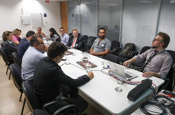 A imagem mostra uma reunião formal em uma sala de conferências. Várias pessoas estão sentadas ao redor de uma mesa branca retangular, participando de uma discussão. Há aproximadamente dez participantes, incluindo homens e mulheres, que parecem estar prestando atenção em uma apresentação feita por um dos integrantes, que está de frente para um laptop e gesticulando enquanto fala. Há copos de vidro com água e jarras sobre a mesa, além de alguns documentos e pastas. No fundo, vê-se divisórias de vidro e portas de correr, e na parede há alguns equipamentos de segurança como extintores.