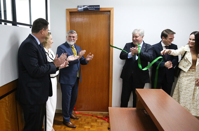 A imagem mostra a cerimônia de inauguração da nova unidade da comarca de Chapecó. Pessoas estão reunidas ao redor de uma porta, e um dos participantes está cortando uma fita verde envolta na porta, enquanto os demais aplaudem.