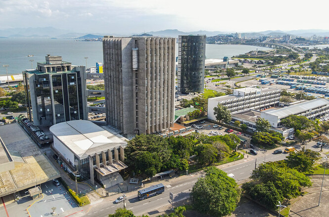 A imagem mostra uma vista aérea de uma área urbana próxima ao mar. No centro, há um conjunto de edifícios altos, com destaque para um prédio de concreto com uma fachada vertical e janelas estreitas. Ao lado, há outro prédio com uma fachada de vidro. Mais ao fundo, é possível ver um edifício cilíndrico de vidro escuro. Há várias árvores e áreas verdes ao redor dos prédios. À direita, há um grande estacionamento com muitos carros. Ao fundo, é possível ver o mar e, mais distante, uma ponte que conecta a cidade a outra área urbana. A paisagem é complementada por montanhas ao longe.