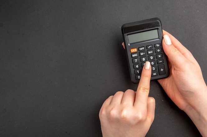 A imagem mostra uma calculadora preta sobre uma superfície escura. Uma pessoa está segurando a calculadora com uma mão e pressionando a tecla "9" com o dedo indicador.