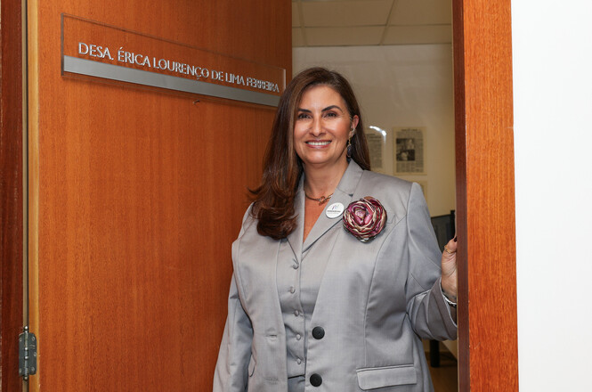 A imagem mostra a desembargadora Érica Lourenço de Lima Ferreira sentada, sorridente, enquanto segura seus óculos. Ela veste blazer cinza com um broche de flor no peito. Alguns documentos estão sobre sua mesa.
