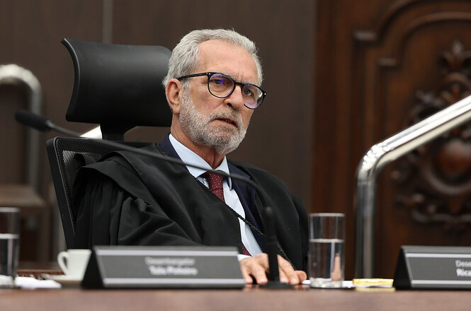 A imagem mostra o desembargador Ricardo Orofino da Luz Fontes. Ele está sentado em uma cadeira de encosto alto, vestindo uma toga. Sobre a mesa à sua frente, há uma placa com seu nome e copos de água.