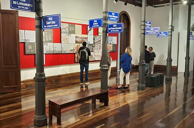 A foto mostra visitantes observando uma exposição no interior do Museu Histórico de Santa Catarina. A exposição é composta por paineis com informações históricas. O chão é de madeira polida, e há bancos e pilares ao longo do espaço.