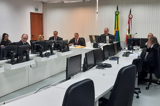 A imagem mostra uma sessão formal em um julgamento, onde diversos profissionais, incluindo juízes e advogados, estão sentados em uma mesa longa com vários computadores.