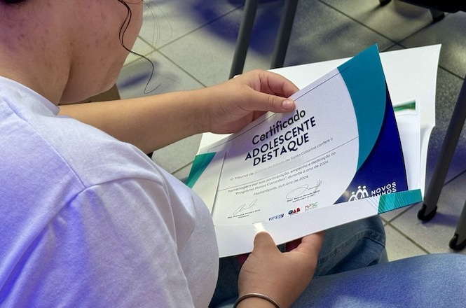 A imagem mostra uma pessoa segurando um certificado. A pessoa está vestindo uma camiseta branca e está sentada em uma cadeira. O certificado está parcialmente visível e tem o título "Certificado Adolescente Destaque". O design do certificado inclui cores como azul e verde, e há logotipos na parte inferior. A pessoa está olhando para o certificado, que parece ser um reconhecimento ou prêmio. O ambiente ao redor parece ser uma sala com piso de azulejos claros e outras cadeiras visíveis ao fundo.